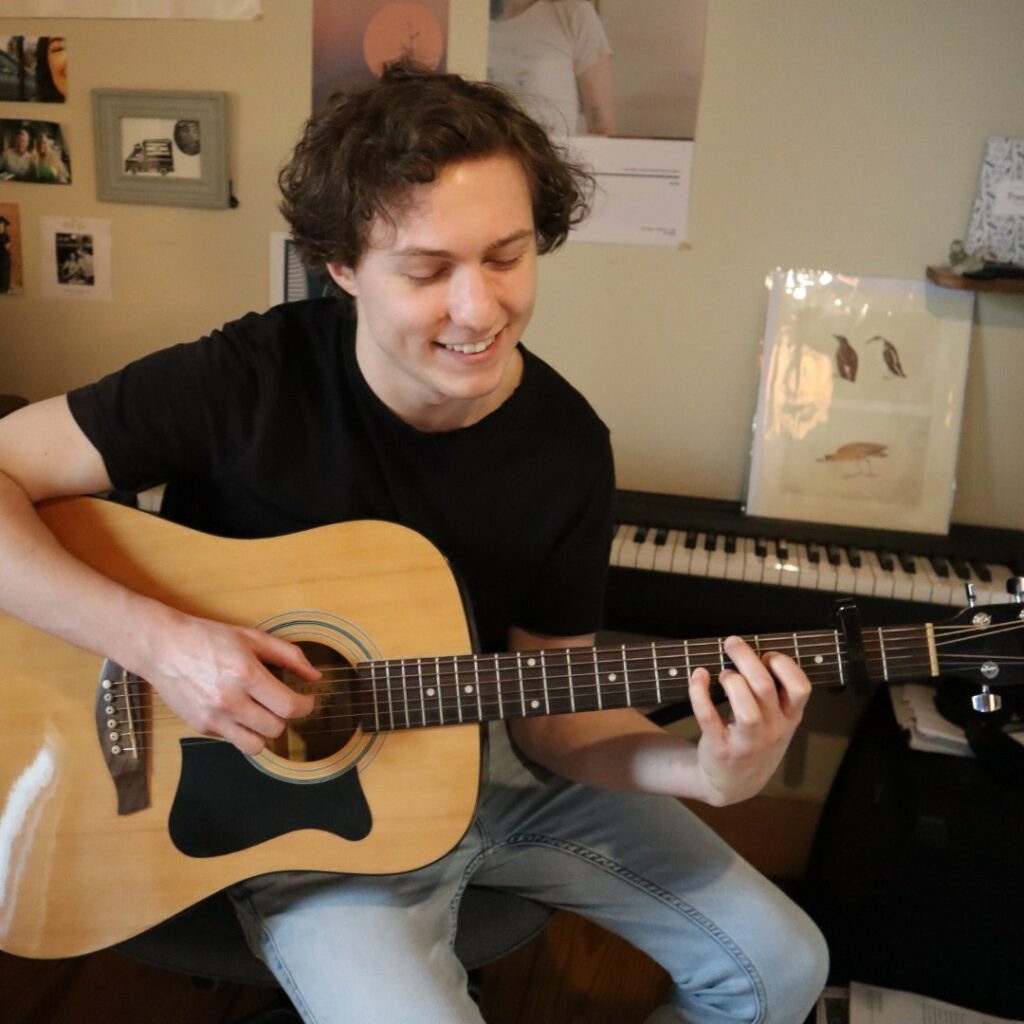 Patrick smiling and playing an acoustic guitar behind a piano.